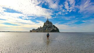 Randonnée à pied dans la baie du Mont-Saint-Michel à marée basse, avec vue sur l’îlot et son abbaye sous un ciel bleu