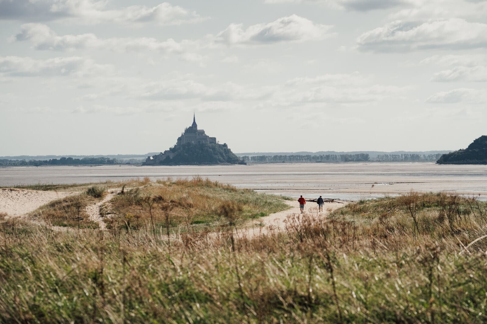 Deux randonneurs marchant à travers les herbus en direction du Mont-Saint-Michel, au cœur de la baie