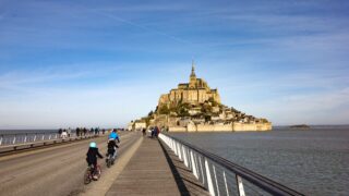 Cyclistes sur la passerelle menant au Mont-Saint-Michel, sous un ciel clair de Normandie