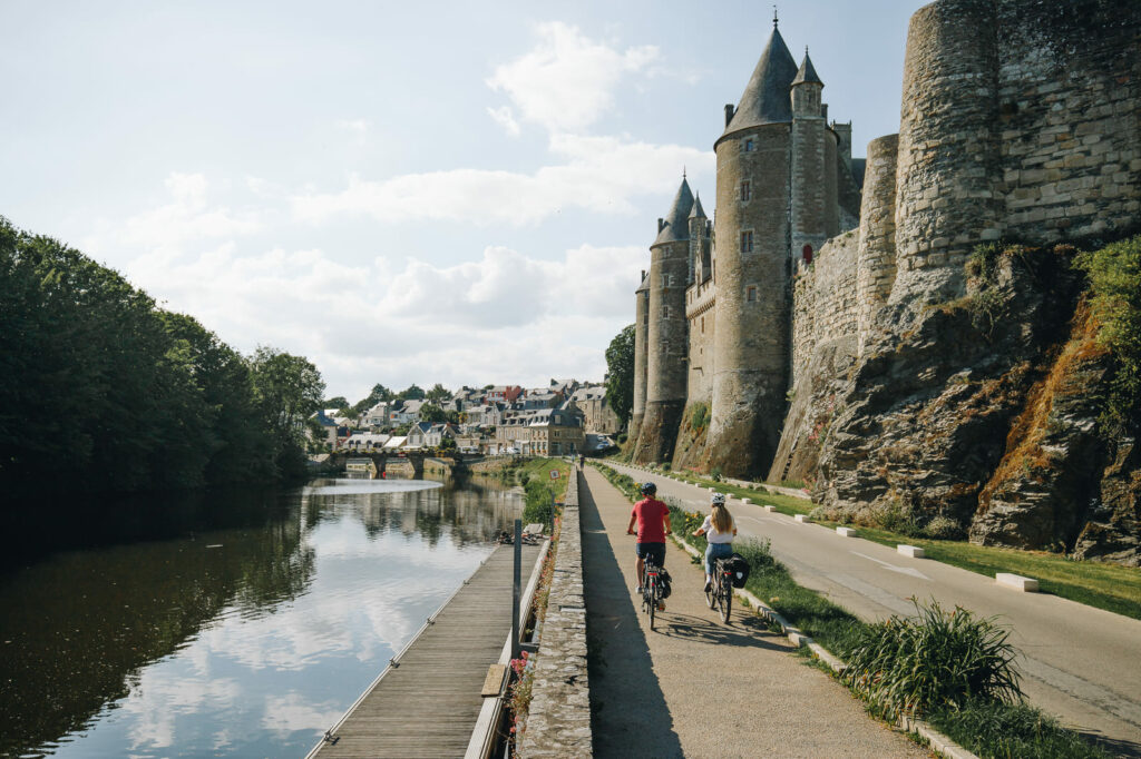 Cyclotouristes longeant le Canal de Nantes à Brest sur la voie verte, avec l'imposant château médiéval de Josselin se dressant sur la rive opposée.