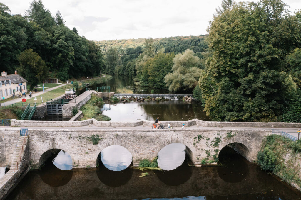 Famille à vélo traversant un vieux pont de pierre au-dessus d'une écluse et de son déversoir sur le Canal de Nantes à Brest.