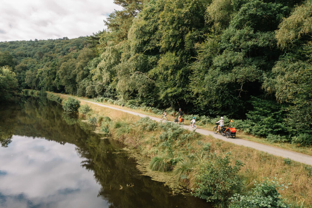 Groupe familial à vélo, incluant une remorque enfant, roulant sur une voie verte bordée d'une forêt dense le long du canal.