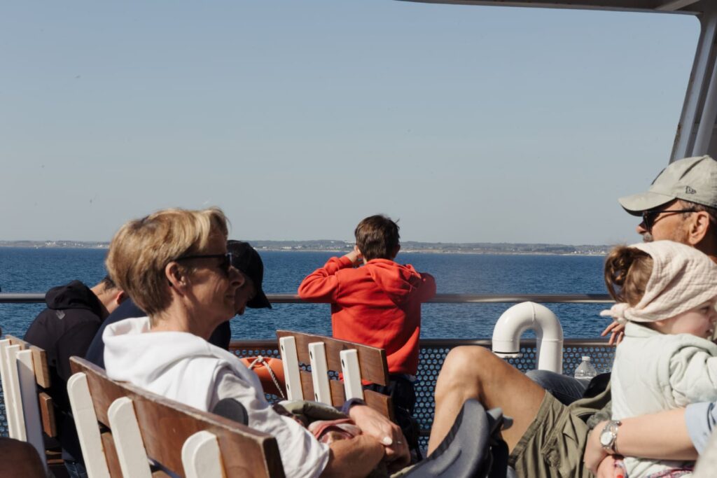 Une famille, dont un enfant avec un sweat à capuche rouge et un bébé dans les bras, regarde la mer et la côte depuis le pont extérieur d'un ferry lors d'une traversée dans le Golfe du Morbihan