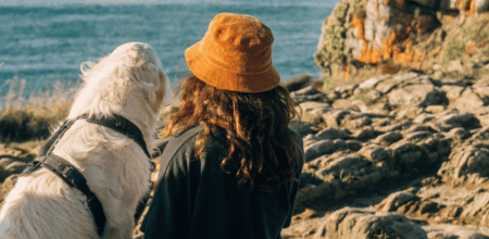 Randonneuse avec un bob orange et son chien blanc équipé d'un harnais, regardant la mer depuis les rochers du GR34.
