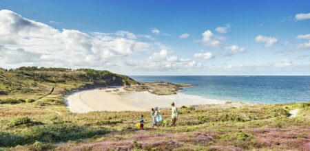 Une famille avec deux jeunes enfants marche sur un sentier à travers la lande en direction d'une plage de sable fin et d'une mer turquoise sous un ciel bleu.